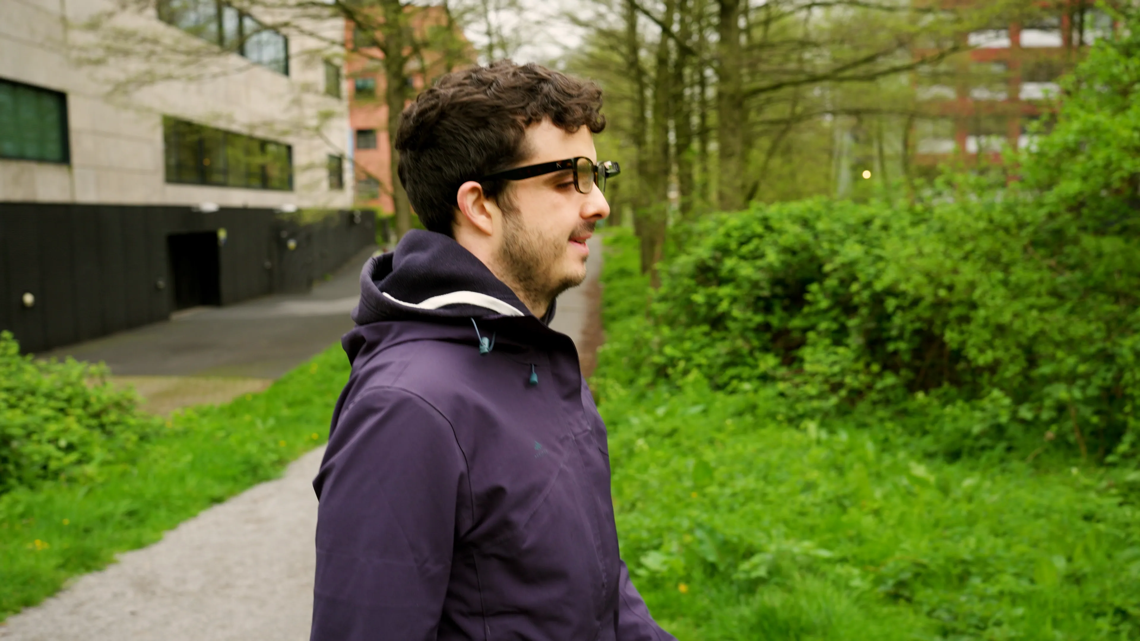 A man with short curly hair and glasses, wearing a dark purple hooded jacket, walks along a path bordered by green grass and bushes. He is in profile view, facing right, with a modern building and trees in the background.